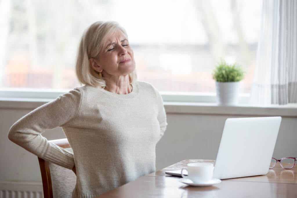 Senior woman in beige sweater at a desk, holding her low back with a painful expression