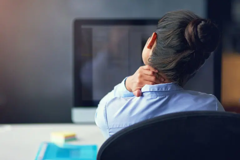 A woman sits at a desk, rubbing her neck and upper back.