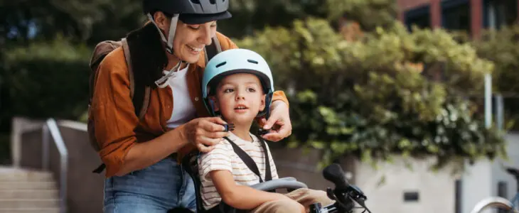 Mother fastening sons' bike helmet on head, carrying him on child bike carrier or seat.